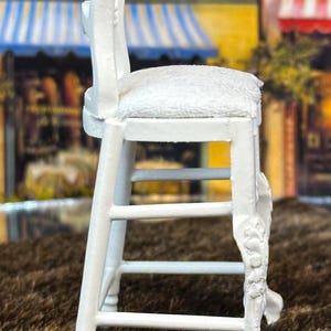 May include: White wooden stool with a padded seat. The stool has a decorative carved design on the legs and back.