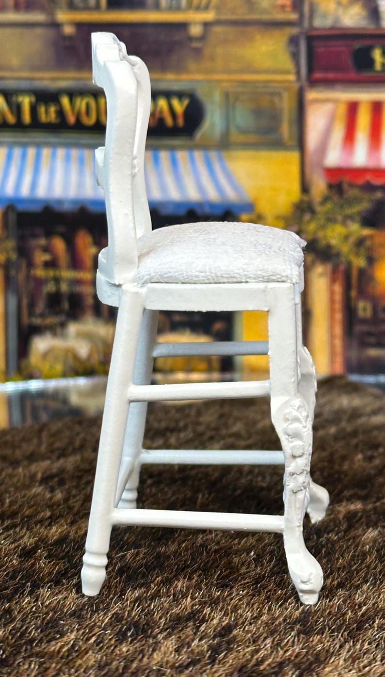 May include: White wooden stool with a padded seat. The stool has a decorative carved design on the legs and back.