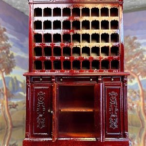 Puede incluir: Un mueble de madera en miniatura, adornado, de color rojo oscuro, con múltiples pequeños huecos y etiquetas numeradas. El mueble tiene dos puertas, una abierta, que revela una estantería. La parte superior del mueble tiene tallas decorativas.