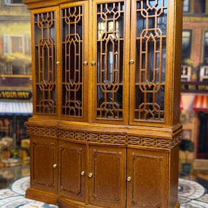 May include: A miniature wooden cabinet with glass-paned doors and decorative fretwork. The cabinet has a dark brown finish, gold-colored hardware, and a detailed design. The cabinet is a dollhouse-sized piece of furniture.