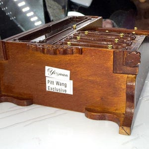 May include: A wooden jewelry box with a rich brown finish. The box features intricate carvings and small gold-colored accents. The label reads "Yiyuan Pitt Wang Exclusive". The box is set on a white surface.