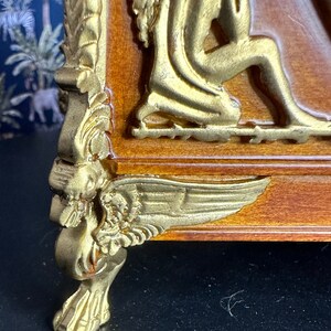 May include: A wooden box with gold accents and a winged cherub design. The box has a decorative foot with a winged cherub design.
