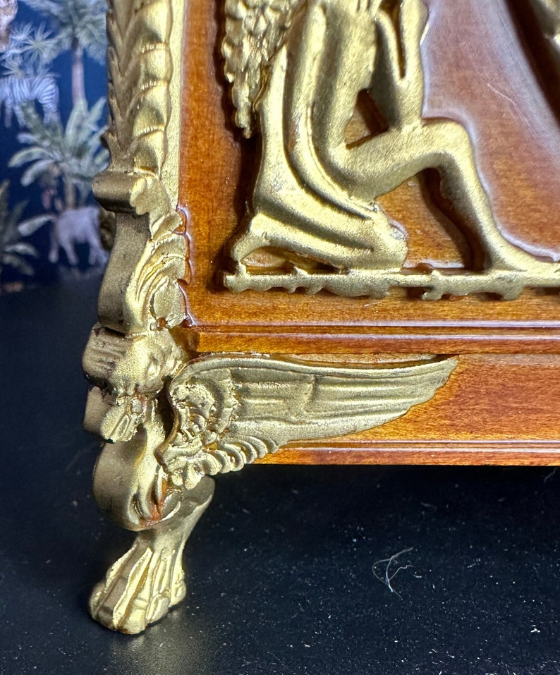 May include: A wooden box with gold accents and a winged cherub design. The box has a decorative foot with a winged cherub design.