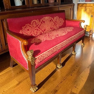 Könnte beinhalten: Eine rot gepolsterte Bank mit einem dekorativen Goldmuster auf Sitz und Rückenlehne. Der Holzrahmen hat verzierte goldene Akzente an den Beinen und Armen. Ein klassisches Möbelstück.
