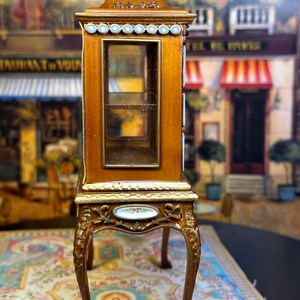 May include: A miniature wooden display cabinet with glass shelves and ornate gold accents. The cabinet has a decorative porcelain plaque on the front and sits on four carved legs.