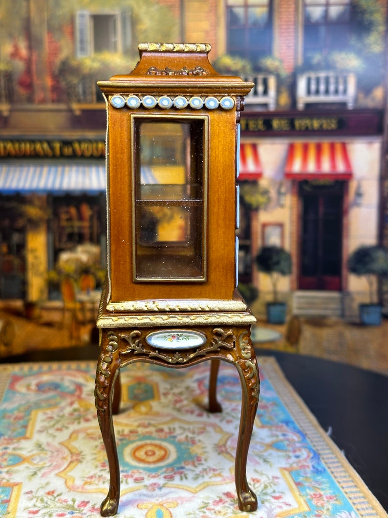 May include: A miniature wooden display cabinet with glass shelves and ornate gold accents. The cabinet has a decorative porcelain plaque on the front and sits on four carved legs.