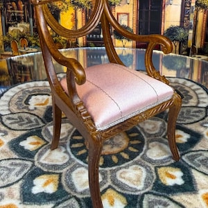 May include: A miniature wooden chair with a pink seat and ornate detailing. The chair has curved arms and legs, and a decorative back. The chair is set on a patterned rug, with a backdrop of a European street scene.