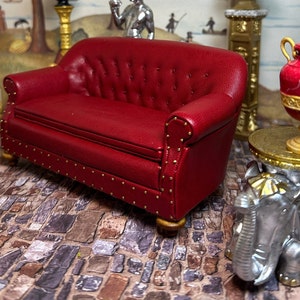 May include: A miniature red leather sofa with button tufting and brass nailhead trim. The sofa has wooden legs and is part of a dollhouse furniture set. A decorative table with an elephant base and a red and gold vase are also visible.