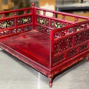 Peut inclure: Un petit lit rouge orné avec des sculptures complexes et des accents floraux. Le cadre du lit comporte une tête de lit et un pied de lit avec des panneaux décoratifs. Le lit est en bois et a une finition brillante. C'est un objet décoratif.