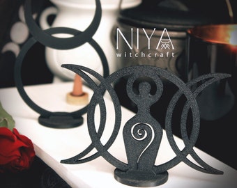 May include: Black wooden triple moon goddess statue with a stylized female figure in the center. The statue is on a white surface with the text "NIYA witchcraft" visible in the background.
