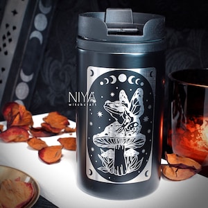 May include: Black stainless steel travel mug with a white design of a frog with wings sitting on a mushroom. The design also includes a crescent moon and stars. The text "NIYA witch craft" is on the mug.