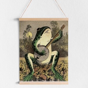May include: A green frog playing a banjo with a brown wooden body and white strings. The frog is sitting on a bed of green and brown leaves. The background is a blurry image of trees and a moon.