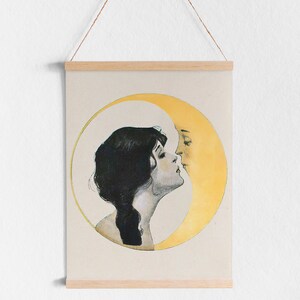 May include: A vintage-style print featuring a woman and a crescent moon in a romantic embrace. The artwork is in soft tones, with the woman's dark hair contrasting against the pale background. The print is held by wooden bars and a hanging string.
