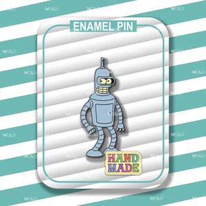 May include: A light blue enamel pin featuring a cartoon robot character with a silver antenna, yellow eyes, and a wide grin. The pin has the text "ENAMEL PIN" at the top and "HAND MADE" at the bottom, set against a striped background.
