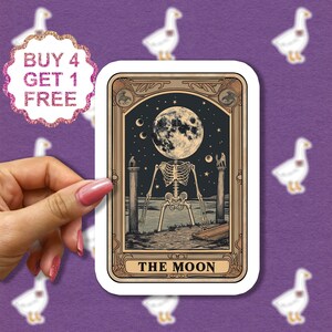 May include: A tarot card with a gold frame and a black and white illustration of a skeleton with a full moon for a head. The card reads "THE MOON" at the bottom.