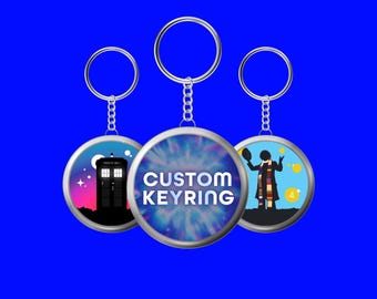 Puede incluir: Tres llaveros circulares de metal con anillas y cadenas plateadas. El llavero central presenta un fondo tie-dye azul y morado con el texto blanco CUSTOM KEYRING. El de la izquierda muestra una cabina azul, el de la derecha una figura.
