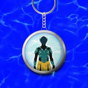 Könnte beinhalten: Ein runder Schlüsselanhänger mit silbernem Rand zeigt eine Silhouette einer Figur in tealfarbener Rüstung und einem gelben Rock vor einem hellblauen Hintergrund. Der Schlüsselanhänger hängt an einer silbernen Kette und einem Ring, vor einem blauen Wasser-ähnlichen Muster.