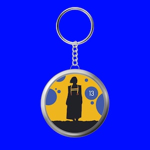 Peut inclure: Un porte-clés rond argenté avec une silhouette d'une personne en robe, sur un fond jaune avec le chiffre 13. Le design est encadré par un bord bleu, le tout sur un fond bleu.