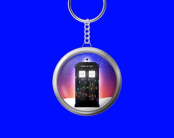 May include: A round silver keychain pendant featuring a black police box with the words "POLICE BOX" on top. The box is decorated with colorful Christmas lights and set against a snowy landscape with a starry sky.