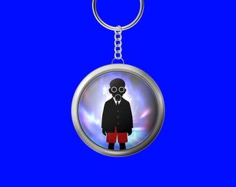 Puede incluir: Un llavero redondo plateado con un colgante que presenta una figura silueteada con máscara de gas, traje negro y pantalones cortos rojos. La figura se encuentra sobre un fondo colorido y arremolinado. El llavero tiene una cadena plateada y un fondo azul.
