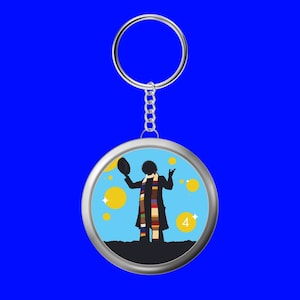 May include: A round silver keychain with a silhouette of a figure in a long coat and scarf, holding a hat. The background is light blue with yellow circles and the number 4. The keychain hangs against a blue background.