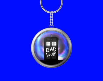 Puede incluir: Un llavero redondo plateado con una imagen en blanco y negro de la TARDIS de Doctor Who, con las palabras "BAD WOLF" en blanco. El fondo es una nebulosa azul y morada.