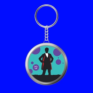 Peut inclure: Un porte-clés argenté avec un pendentif rond représentant la silhouette d'une personne portant un manteau doublé de rouge. Le fond est turquoise avec des cercles violets et le chiffre 12. Le porte-clés a une chaîne et un anneau argentés.