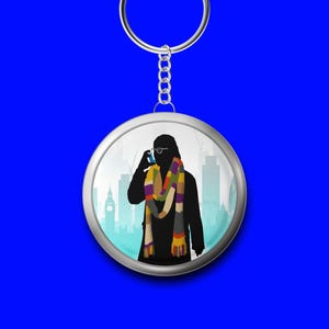 Peut inclure: Un porte-clés rond avec une bordure argentée et un fond bleu. Il présente une silhouette d'une personne portant une écharpe colorée et tenant un téléphone, avec un paysage urbain en arrière-plan.