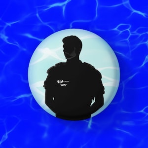 Könnte beinhalten: Ein runder Button mit einer schwarzen Silhouette einer Person in Uniform, vor einem hellblauen Hintergrund. Die Person befindet sich in einer hellblauen Kugel, mit dem Wort "UNIT" und einem Logo auf der Uniform. Der Hintergrund ist ein blaues Wassermuster.