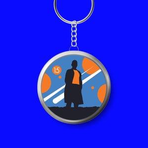 May include: A circular keyring featuring a graphic illustration of a silhouette standing against a blue background with orange circles, white diagonal streaks, and the number 15 in a small orange circle. The keyring has a silver metal ring.