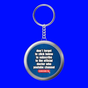 Peut inclure: Un porte-clés circulaire avec un bord en métal argenté et un centre bleu. Le texte indique : don't forget to click below to subscribe to the official doctor who youtube channel. Sous le texte se trouve un bouton rectangulaire rouge avec le mot SUBSCRIBE.