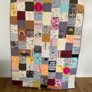 May include: A rectangular memory quilt constructed from various fabric squares featuring diverse patterns, colours, and printed text including "DADDY + ME BEST MATES FOREVER," "mummy's coffee date," "DAD IS MY SUPER HERO," and "WILD AT HEART" with a grey border.