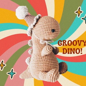 Puede incluir: Un juguete de dinosaurio de crochet que lleva un gorro de fiesta con un pompón blanco. El dinosaurio es de color canela y tiene un ojo negro. El fondo es un patrón de espiral colorido con el texto "GROOVY DINO!" en rojo.