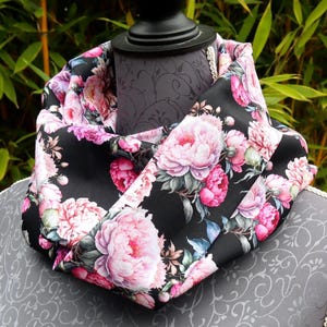 May include: A black infinity scarf with a vibrant floral pattern of pink peonies and green leaves. The scarf is draped over a mannequin, showcasing the design and fabric. The background is a lush green.