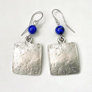 May include: A pair of silver earrings featuring a textured square pendant and a deep blue bead. The earrings have a simple hook design and are set against a white backdrop. These are a type of jewellery.