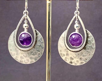 May include: Silver earrings with a hammered texture and a teardrop design. Each earring features a vibrant purple amethyst gemstone. The earrings are displayed on a gold-coloured stand against a dark background.
