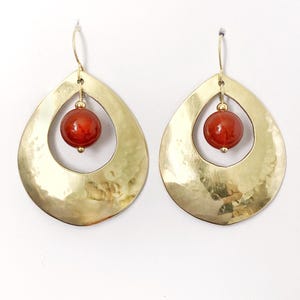 May include: A pair of gold-toned teardrop-shaped earrings featuring a hammered metal texture. Each earring has a central circular opening containing a single, smooth, round, deep orange-red bead suspended from a thin gold-toned wire hook.