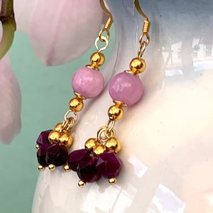 A pair of beaded bohemian earrings with pink & purple gemstone, are shown hanging from a white vase against a dark background.