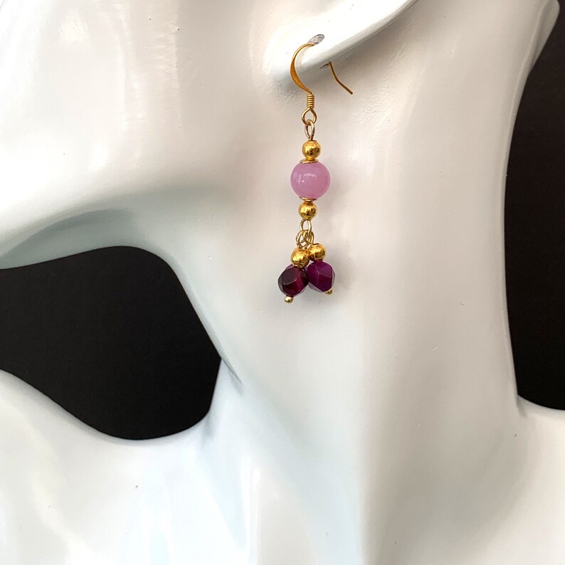 A beaded bohemian earring with pink & purple gemstone, is shown hanging on a white head manequin, against a warm brown/ black background. By Earrings of Gemstone.