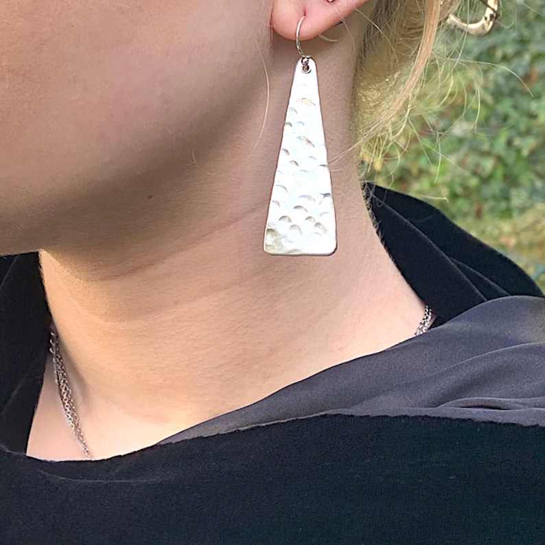 A pair of long triangle geometric hammered earrings with sterling silver hooks is shown on a human model in front of a green outdoor background. By Earrings Of Gemstone.