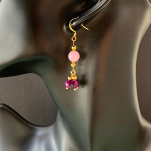 A beaded bohemian earring with pink & purple gemstone, is shown hanging on a black head manequin, against a warm brown/ black background. By Earrings of Gemstone.