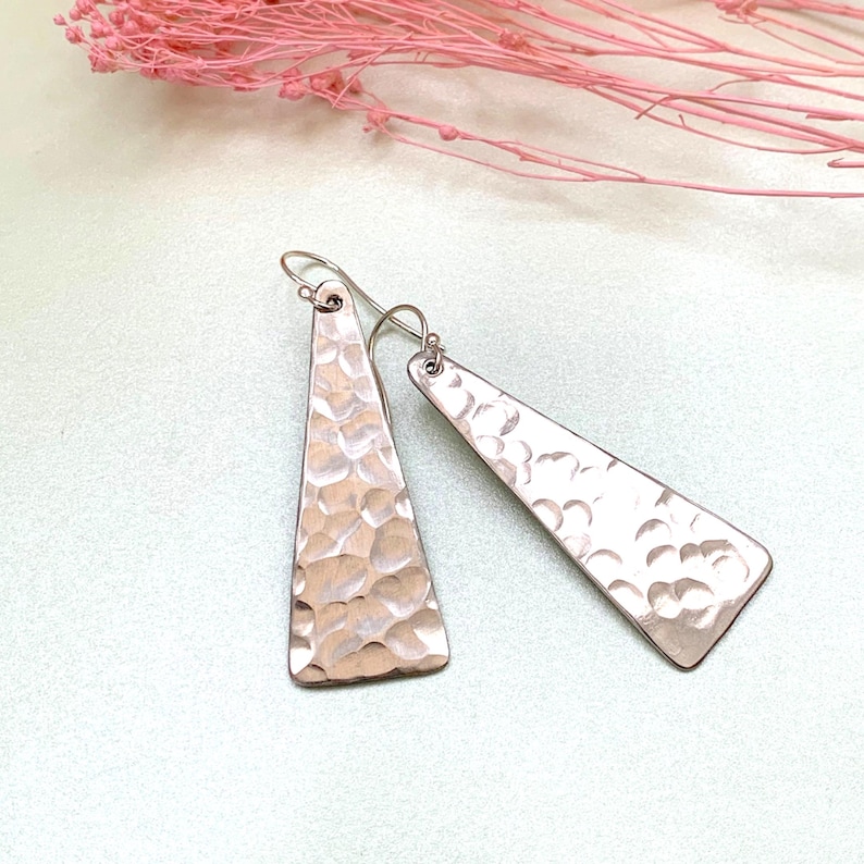 A pair of long triangle geometric hammered earrings with sterling silver hooks is shown on a light green background with delicate pink flower sprigs. By Earrings Of Gemstone.