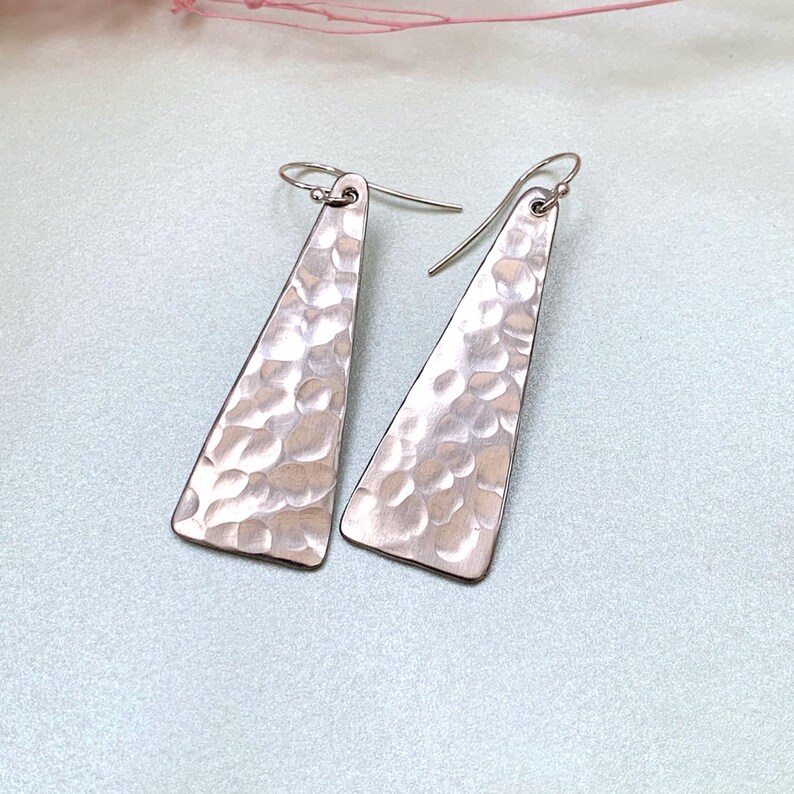 A pair of long triangle geometric hammered earrings with sterling silver hooks is shown on a light green background with delicate pink flower sprigs. By Earrings Of Gemstone.