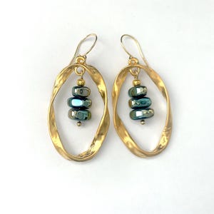 May include: A pair of gold-toned, hammered-look oval earrings. Each earring features a stack of four iridescent teal beads suspended within the oval. The earrings have a simple hook closure.