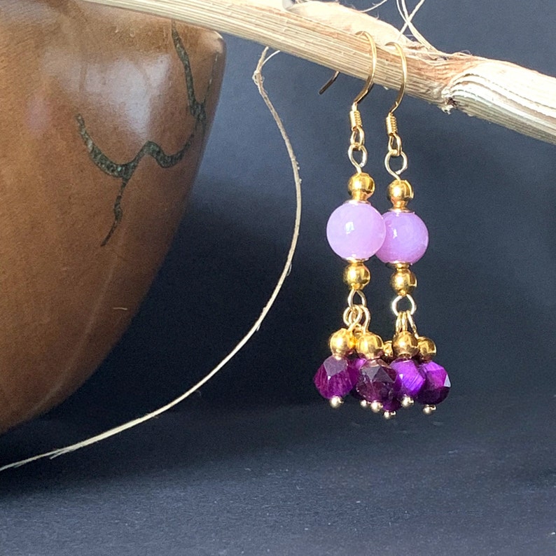 A pair of beaded bohemian earrings with pink & purple gemstone, are shown hanging from the rim of a dried palm leaf stem, against a warm brown background.