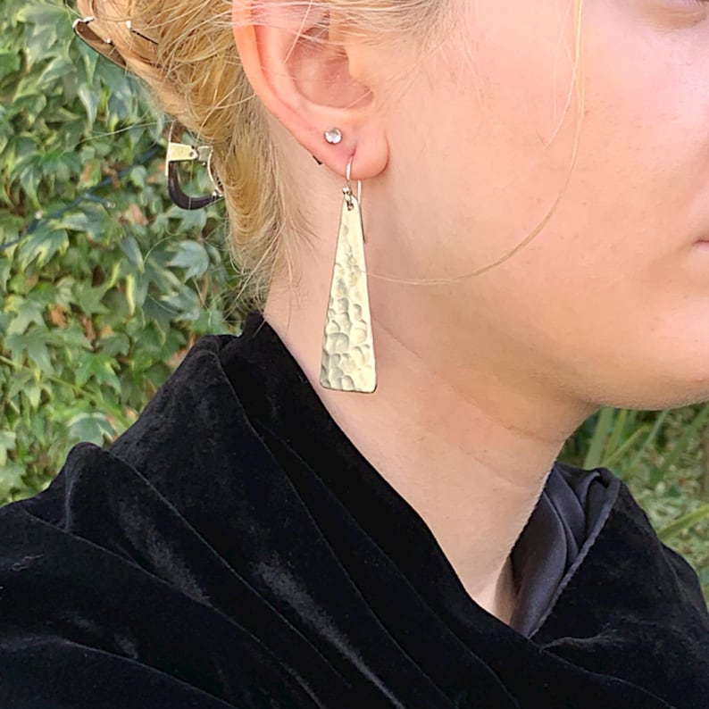 A pair of long triangle geometric hammered earrings with sterling silver hooks is shown on a human model in front of a green outdoor background. By Earrings Of Gemstone.