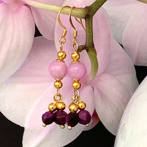 A pair of beaded bohemian earrings with pink & purple gemstone, are shown hanging from a pink orchid flower, against a warm brown background.