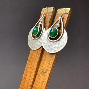 May include: Silver earrings with a hammered finish and a teardrop shape. Each earring has a bright green stone in a wire setting, providing a splash of colour. The earrings are shown on a wooden ruler.