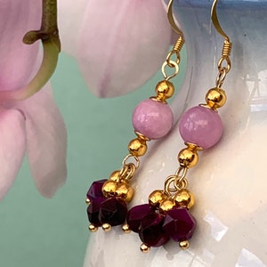 A pair of beaded bohemian earrings with pink & purple gemstone, are shown hanging from a white vase against a dark background.