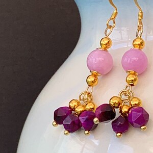 A pair of beaded bohemian earrings with pink & purple gemstone, are shown hanging from a white vase against a dark background.